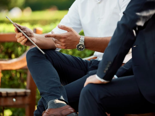 Two men in business attire sitting on a bench, engaged in conversation in an outdoor setting.