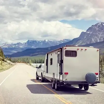A white truck tows a camper along a scenic mountain road, bordered by forests and majestic peaks under a cloudy sky, conveying freedom and adventure.