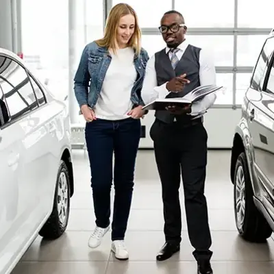 A woman and salesman are in a car showroom, discussing options. She is smiling, and he is holding a booklet. Bright, modern setting with cars displayed.