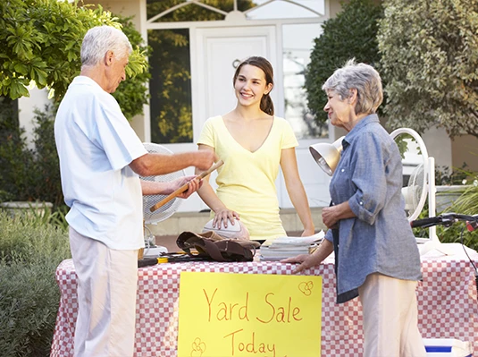 Elderly couple and young woman at a yard sale table outdoors. The woman smiles behind a sign reading "Yard Sale Today." Warm, friendly atmosphere.