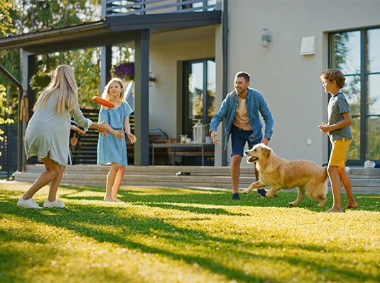 A family of four and a dog play with a frisbee on a sunny lawn. The scene exudes joy and togetherness, with a modern house in the background.