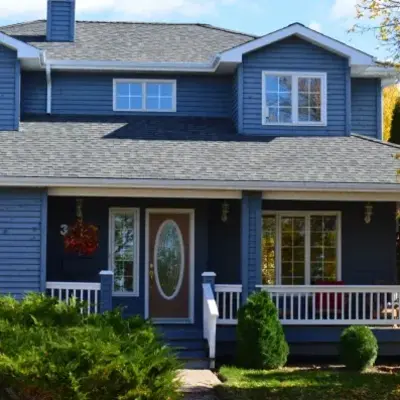 A charming two-story blue house with white trim, features a spacious porch, oval glass door, and autumn foliage, creating a welcoming, cozy atmosphere.