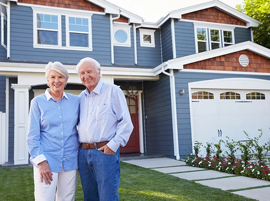 An elderly couple stands in front of a blue two-story house with white trim, smiling warmly. The setting is bright and welcoming, suggesting comfort and happiness.