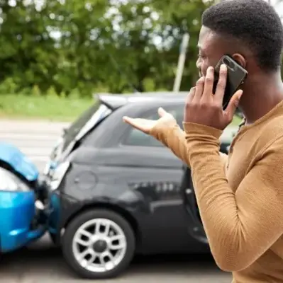 A man stands beside a damaged car, talking on his cell phone with a concerned expression.