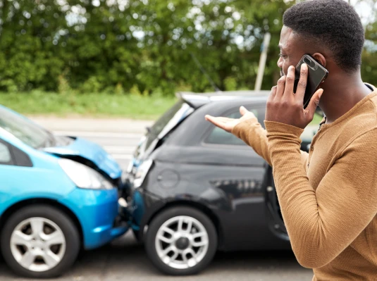 A man stands beside a damaged car, talking on his cell phone with a concerned expression.