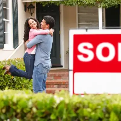 A joyful couple embraces in front of their new home, with a large "SOLD" sign in the foreground. The scene conveys happiness and new beginnings.