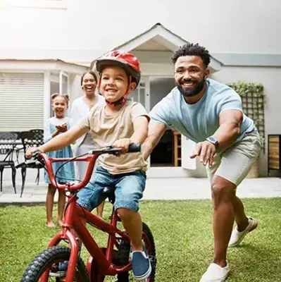 A joyful family scene in a backyard. A child rides a bike with a red helmet, while an adult, smiling, guides them. Two others watch happily in the background.