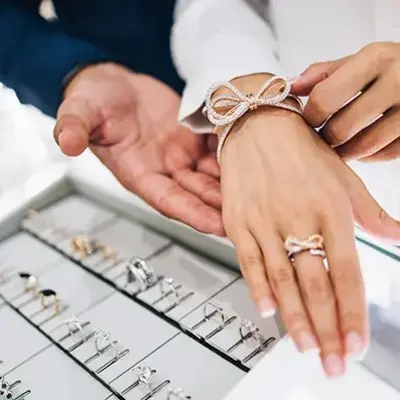Close-up of a jewelry store display. A hand tries on an elegant bow-shaped bracelet and ring, with a salesperson’s hand assisting, evoking elegance and luxury.