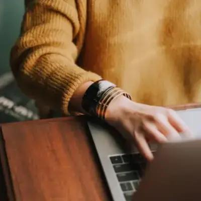 A person in a mustard sweater types on a laptop at a wooden desk, wearing multiple bracelets. The scene conveys focus and productivity.