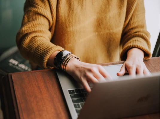 A person in a mustard sweater types on a laptop at a wooden desk, wearing multiple bracelets. The scene conveys focus and productivity.