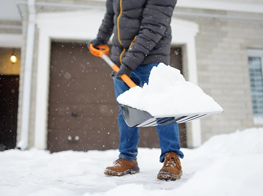 Person in winter clothing shoveling snow on a driveway. They wear a gray jacket, blue jeans, and brown boots. Snowflakes fall gently, creating a chilly atmosphere.