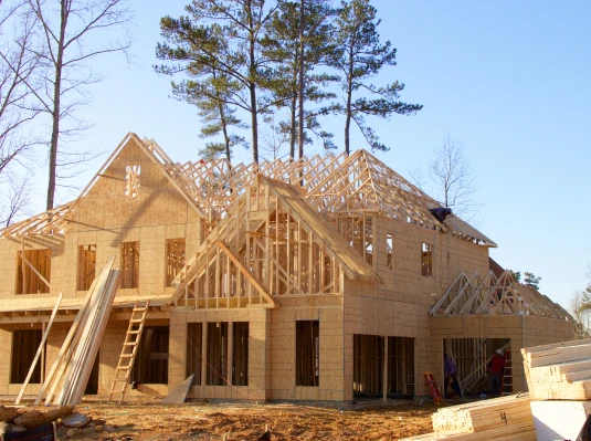 A two-story house under construction with exposed wooden framing and trusses, surrounded by trees on a clear day, conveying progress and potential.
