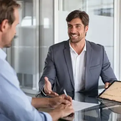 Three people in an office meeting; one man in a suit is showing a tablet with charts, while another takes notes.