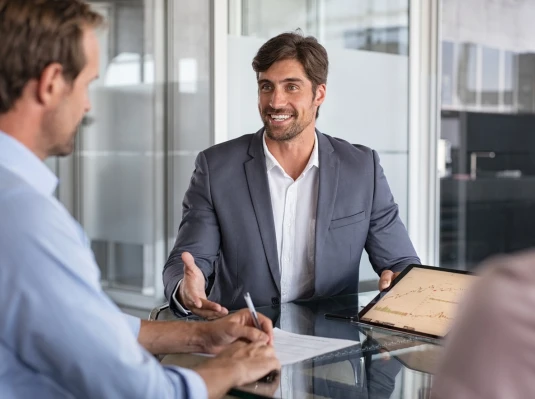 Three people in an office meeting; one man in a suit is showing a tablet with charts, while another takes notes.