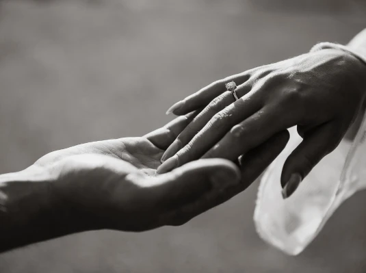 A close-up black and white photo shows two hands reaching towards each other. The hand on the right has a diamond ring, symbolizing love and connection.