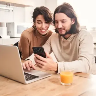 A man and woman are engaged in conversation while looking at a laptop and a mobile phone together.