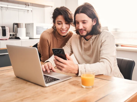 A man and woman are engaged in conversation while looking at a laptop and a mobile phone together.