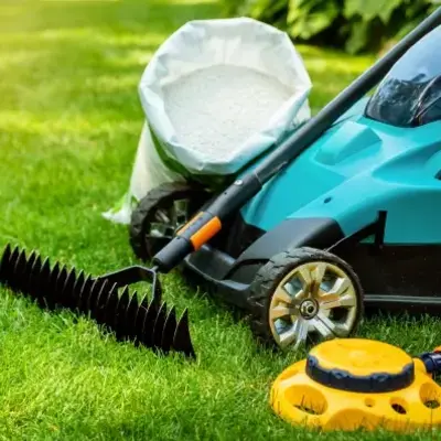 A blue lawn mower, a black rake, a bag of white fertilizer, and a yellow sprinkler lie on lush green grass, conveying a gardening theme.