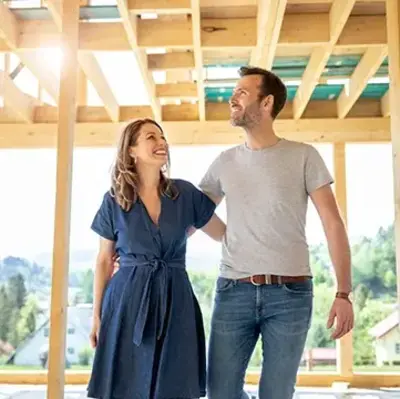 A couple happily walks through a sunlit, unfinished wooden house frame, smiling at each other. The background reveals a scenic, tree-filled landscape.