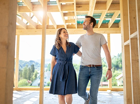 A couple happily walks through a sunlit, unfinished wooden house frame, smiling at each other. The background reveals a scenic, tree-filled landscape.