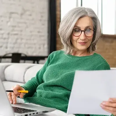 Elderly woman in green sweater sitting on a couch, holding a paper and pen, focused. Open laptop beside her; cozy, modern interior.