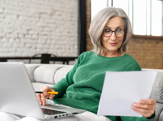 Elderly woman in green sweater sitting on a couch, holding a paper and pen, focused. Open laptop beside her; cozy, modern interior.
