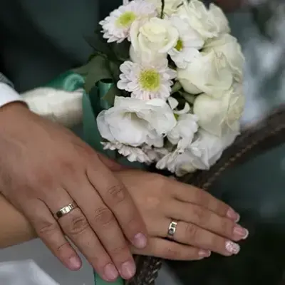 Hands with wedding rings resting on a white dress, holding a bouquet of white roses and daisies. The image conveys a sense of love and unity.