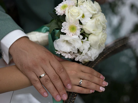 Hands with wedding rings resting on a white dress, holding a bouquet of white roses and daisies. The image conveys a sense of love and unity.