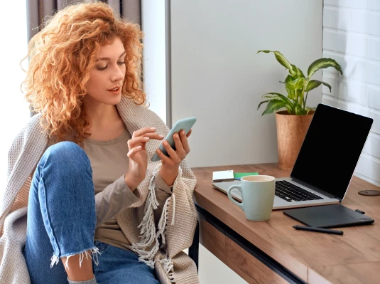 A woman seated at a desk, working on a laptop with a smartphone beside her.
