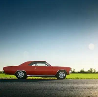 Red vintage car parked on a rural road under a clear blue sky. Sunlight brightens the green field in the background, conveying a serene, nostalgic mood.