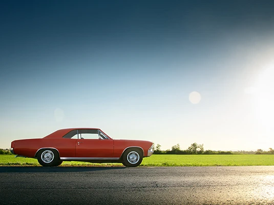 Red vintage car parked on a rural road under a clear blue sky. Sunlight brightens the green field in the background, conveying a serene, nostalgic mood.