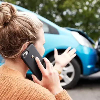 A woman in a sweater is on the phone, gesturing toward a minor collision between two cars, one blue and one black. The mood appears tense and urgent.