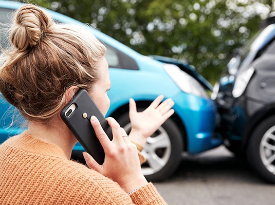 A woman in a sweater is on the phone, gesturing toward a minor collision between two cars, one blue and one black. The mood appears tense and urgent.