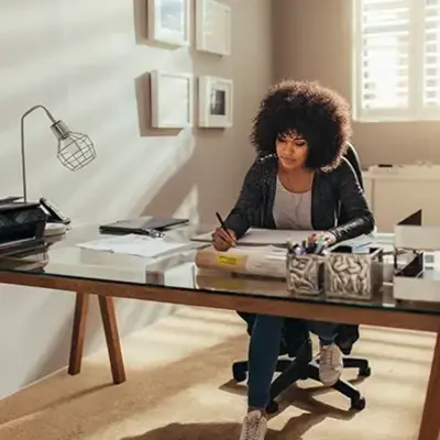 Woman focused on work at a modern home office desk with glass tabletop. Sunlight filters through blinds, creating a warm, serene atmosphere.