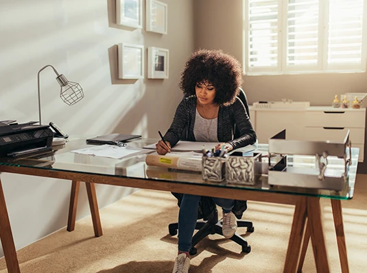 Woman focused on work at a modern home office desk with glass tabletop. Sunlight filters through blinds, creating a warm, serene atmosphere.