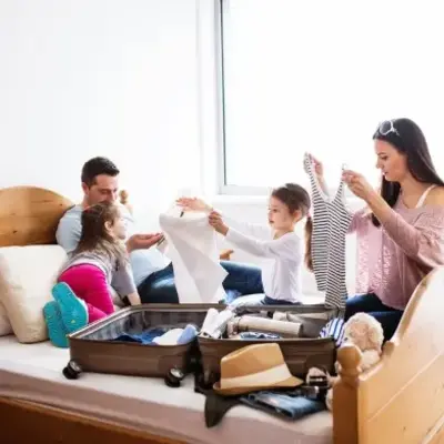 A family of four joyfully packs a suitcase on a bed. Parents and two young girls fold clothes. The room is bright with natural light, creating a cheerful atmosphere.