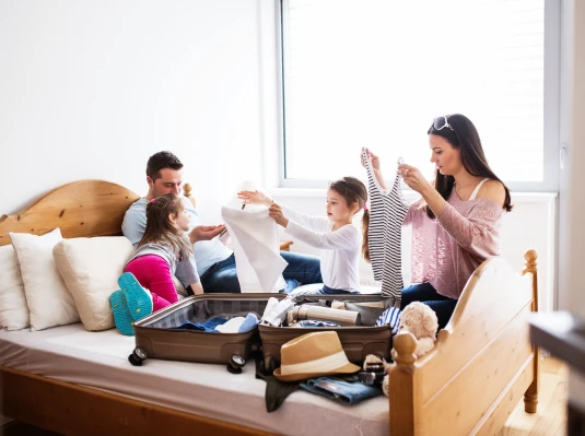 A family of four joyfully packs a suitcase on a bed. Parents and two young girls fold clothes. The room is bright with natural light, creating a cheerful atmosphere.