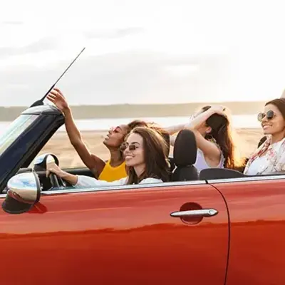 Four women are joyfully riding in a red convertible by the beach, with their hair blowing in the wind. They wear sunglasses, enjoying a carefree, sunny day.