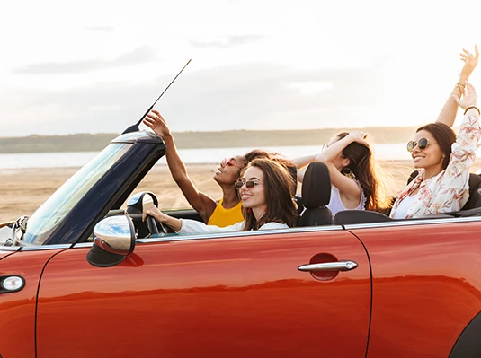 Four women are joyfully riding in a red convertible by the beach, with their hair blowing in the wind. They wear sunglasses, enjoying a carefree, sunny day.