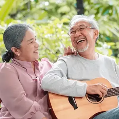 Elderly couple sitting on a bench, smiling joyfully. The man plays an acoustic guitar while the woman looks at him affectionately. Lush greenery in the background.