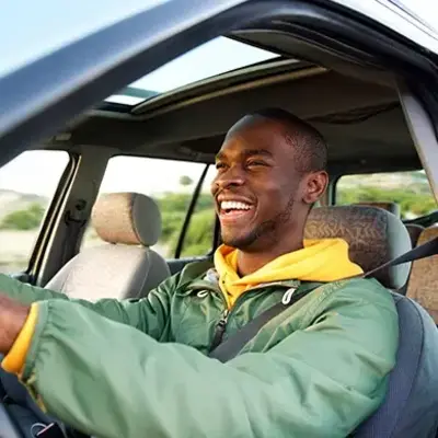 A man in a green jacket smiles joyfully while driving a car with a sunroof. The bright, open landscape outside suggests a pleasant day.