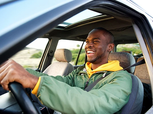 A man in a green jacket smiles joyfully while driving a car with a sunroof. The bright, open landscape outside suggests a pleasant day.