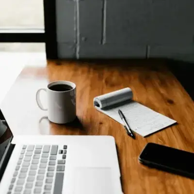 A laptop, a cup of coffee, and a notepad arranged on a wooden desk, creating a cozy workspace atmosphere.