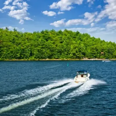 A white speedboat cruises across a blue lake, leaving a foamy wake. The scene is calm, with a lush green forest under a bright sky with scattered clouds.