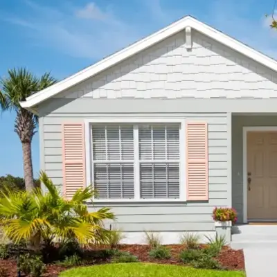 Charming small house with light blue siding, pink shutters, and a tan door. Lush lawn, vibrant flowers, palm tree, and bright blue sky evoke a cheerful, welcoming vibe.