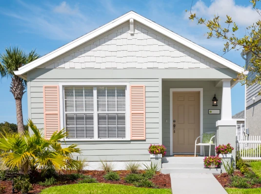 Charming small house with light blue siding, pink shutters, and a tan door. Lush lawn, vibrant flowers, palm tree, and bright blue sky evoke a cheerful, welcoming vibe.