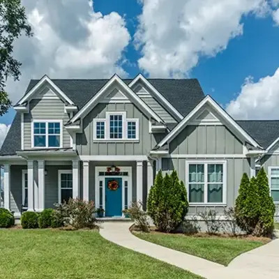 A classic gray house with white trim, a blue door, and a welcoming wreath. It's surrounded by green bushes and trees, under a bright blue sky.