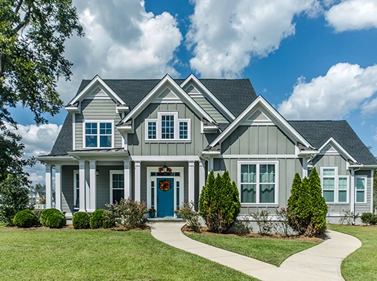 A classic gray house with white trim, a blue door, and a welcoming wreath. It's surrounded by green bushes and trees, under a bright blue sky.