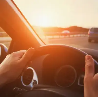 Hands grip a steering wheel, driving on a sunny highway. Sunlight streams in, casting a warm glow over the road with scattered cars ahead.