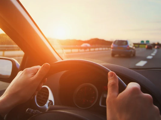 Hands grip a steering wheel, driving on a sunny highway. Sunlight streams in, casting a warm glow over the road with scattered cars ahead.